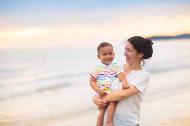 Anne ve bebek gün batımında tropik plajda. Denizde yürüyen ve oynayan küçük bir çocukla Asyalı bir kadın. Genç anne ve küçük çocuk okyanus kıyısında yürüyorlar. Çocuklarla seyahat et. Küçük çocukla tatil..