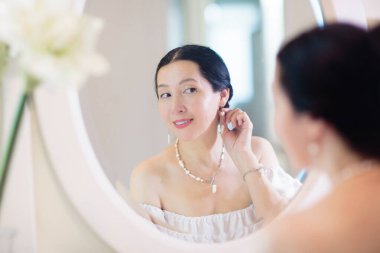 Genç Asyalı kadın aynaya bakıyor. Cilt bakımı rutini. Randevu gecesi ya da hafta sonu sabahında krem süren gecelikli güzel bir kadın. Deri temizleme ve tedavi. Mücevher ve inciler.. 