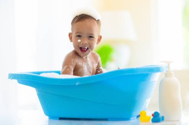 Bebek banyoda. Anne bebeğini yıkıyor. Beyaz güneşli banyoda sabun köpüğüyle oynayan tatlı küçük bebek. Su eğlencesi. Küçük çocuklar için saç ve cilt bakımı. Şampuan ve vücut losyonu. Banyo oyuncakları.