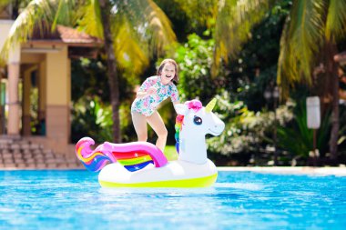 Tek boynuzlu at üzerindeki çocuk tropik tatil köylerinin havuzunda yüzüyor. Çocuklar yüzer ve şişme su oyuncaklarıyla oynarlar. Egzotik adada yaz tatilinde pembe midilli yüzüğünde oynayan küçük bir kız. Kumsal eğlencesi.