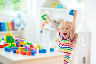 Çocuk renkli oyuncak bloklarıyla oynuyor. Küçük çocuk oyuncaklardan kule inşa ediyor. Küçük çocuklar için eğitici ve yaratıcı oyuncaklar ve oyunlar. Gökkuşağı tuğlalı beyaz yatak odasındaki bebek. Evdeki çocuk.