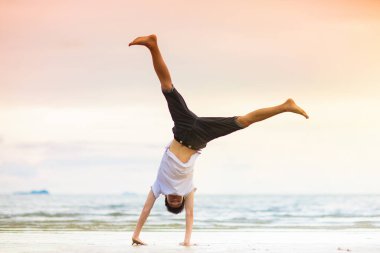Gençler jimnastik yapıyor. Gün batımında plaj yoga. Genç çocuk egzersiz yapıyor. Tekerlek ve amuda kalkma poz verme eğitimi. Aktif genç bir yetişkin için spor. Kol dengesi Asana. Okul çocuk sporları.