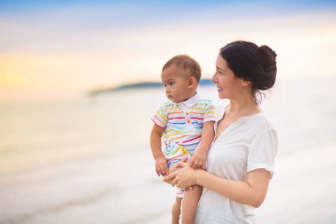 Anne ve bebek gün batımında tropik plajda. Denizde yürüyen ve oynayan küçük bir çocukla Asyalı bir kadın. Genç anne ve küçük çocuk okyanus kıyısında yürüyorlar. Çocuklarla seyahat et. Küçük çocukla tatil..