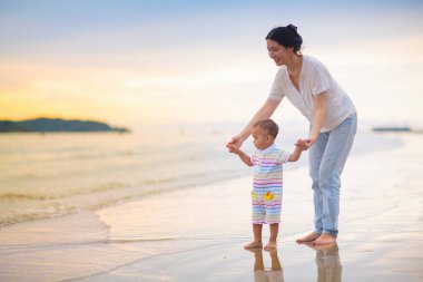 Anne ve bebek gün batımında tropik plajda. Denizde yürüyen ve oynayan küçük bir çocukla Asyalı bir kadın. Genç anne ve küçük çocuk okyanus kıyısında yürüyorlar. Çocuklarla seyahat et. Küçük çocukla tatil..