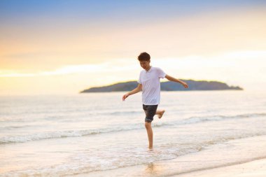 Okyanus sahilinde oynayan bir genç. Asyalı genç gün batımında dalgalara atlıyor. Aile için deniz tatili. Çocuk yaz tatilinde egzotik adada koşuyor..