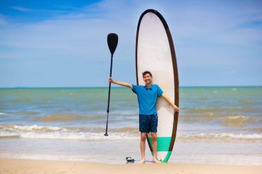 Çocuk ayağa kalkmış kürek çekiyor. Çocuklar için su eğlencesi ve plaj sporu. Sörf tahtasındaki küçük çocuk. Tropikal adada yaz tatili için sağlıklı açık hava sporları. Tatil aktivitesi. Spor yapan sörfçü.