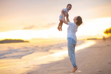 Anne ve bebek gün batımında tropik plajda. Denizde yürüyen ve oynayan küçük bir çocukla Asyalı bir kadın. Genç anne ve küçük çocuk okyanus kıyısında yürüyorlar. Çocuklarla seyahat et. Küçük çocukla tatil..