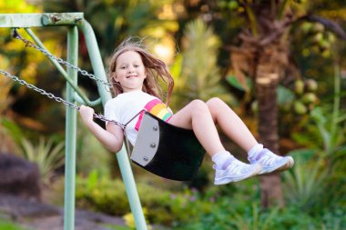 Çocuk oyun parkında güneşli yaz gününde bir parkta sallanan. Çocuklar salıncak. Zemin okul veya anaokulu bahçesinde ve oyun. Küçük kız yüksekten salıncakta havada uçuyor. Yaz açık hava etkinliği. Oynayan çocuk.