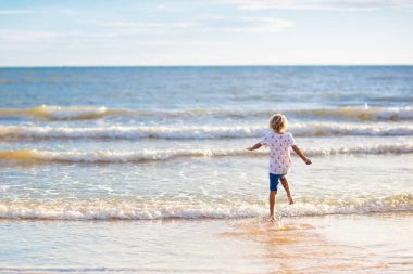 Tropik plajda oynayan bir çocuk. Deniz kıyısında elinde kova ve kürek olan küçük bir çocuk. Aile yaz tatili. Çocuk kumdan kale yapıyor. Çocuklar deniz kabuğu arar ve oynarlar. Çocuklar için su ve kum eğlencesi. 