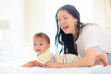 Anne ve çocuk beyaz bir yatakta. Bebek bezli Asyalı anne ve çocuk güneşli yatak odasında oynuyorlar. Ebeveyn ve küçük çocuk evde dinleniyor. Aile birlikte eğleniyor. Bebek odası için yatak ve tekstil.