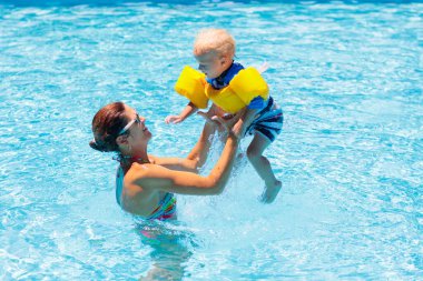 Anne ve bebek tropikal tatil köylerinin havuzunda yüzüyorlar. Çocuk yüzmeyi öğreniyor. Anne ve çocuk suda oynuyorlar. Egzotik bir yerde aile tatili. Çocuklar için aktif ve sağlıklı spor.