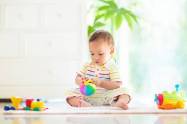 Yakışıklı Asyalı bebek, beyaz güneşli yatak odasında renkli oyuncaklarla sürünmeyi ve oynamayı öğreniyor. Oyun paspasında emekleyen sevimli gülen çocuk. Küçük çocuklar için çocuk odası, giysi ve oyuncaklar..