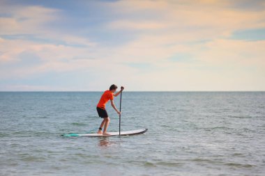 Ayağa kalkmış kürek çeken genç adam. Su eğlencesi ve plaj sporu. Tropikal adada yaz tatili için sağlıklı açık hava sporları. Tatil aktivitesi. Formda bir genç çocuk eğitimi. Spor yapan sörfçü.