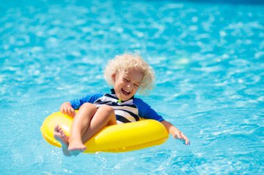 Yüzme havuzunda şişme yüzük takan çocuk. Sarışın kıvırcık çocuk yüzmeyi ve açık havuza dalmayı öğreniyor. Çocuklarla yüzmek. Çocuklar için sağlıklı yaz sporları aktivitesi. Güneş koruması. Su eğlencesi