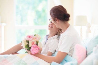 Çocuk annesine yatakta hediye veriyor. Anneler günü hediyesi. Çiçek buketli küçük çocuk beyaz güneşli yatak odasında annesine sarılıyor. Doğum günü kutlaması. Anneler gününde yatakta kahvaltı. Aile sevgisi.