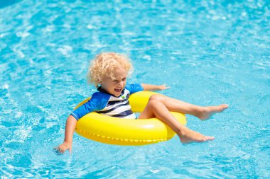 Yüzme havuzunda şişme yüzük takan çocuk. Sarışın kıvırcık çocuk yüzmeyi ve açık havuza dalmayı öğreniyor. Çocuklarla yüzmek. Çocuklar için sağlıklı yaz sporları aktivitesi. Güneş koruması. Su eğlencesi