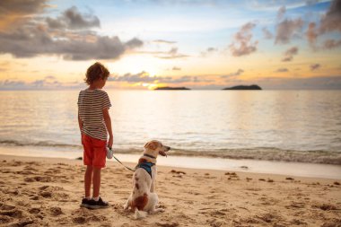 Çocuk ve köpek gün batımında tropik plajda oynuyorlar. Küçük çocuk köpeğiyle koşuyor. Aile yaz tatili tarafı. Okyanusta gün doğumu. Çocuklarla ve evcil hayvanlarla seyahat et. Çocuklar ve hayvanlarla deniz tatili. 