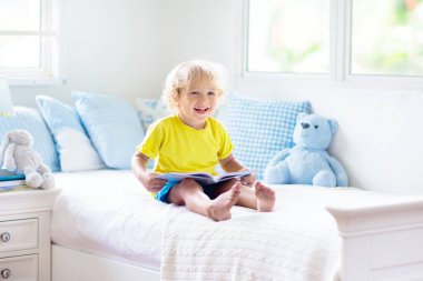 Beyaz güneşli bir odada pencereli bir çocuk yatakta oynuyor. Çocuk odası ve iç tasarım. Erkek bebek evde. Çocuk yuvası için yatak ve tekstil. Oyuncağı ve kitabı olan bir çocuk. Uyku ve uyku zamanı..