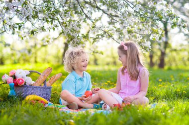 Bahar Parkı 'nda çiçek açan kiraz ağaçlarıyla aile pikniği. Çocuklar dışarıda meyve ve ekmek yiyorlar. Çiçekli elma bahçesinde piknik sepetiyle battaniyenin üzerinde oturuyorlar. Çocuklar için sağlıklı beslenme.