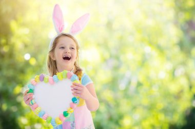 Holding bir kalp kurulu renkli Paskalya yumurtaları ile tavşan kulakları mutlu küçük kız. Çocukları Paskalya kutlamak. Çocuk Paskalya yumurtası avına eğleniyor. Pastel bunny afiş ve çiçekler. Senin metin için yer.