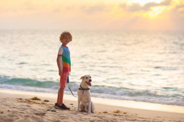 Çocuk ve köpek gün batımında tropik plajda oynuyorlar. Küçük çocuk köpeğiyle koşuyor. Aile yaz tatili tarafı. Okyanusta gün doğumu. Çocuklarla ve evcil hayvanlarla seyahat et. Çocuklar ve hayvanlarla deniz tatili. 