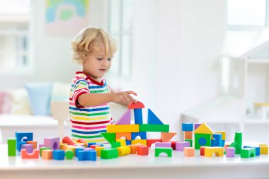 Çocuk renkli oyuncak bloklarıyla oynuyor. Küçük çocuk oyuncaklardan kule inşa ediyor. Küçük çocuklar için eğitici ve yaratıcı oyuncaklar ve oyunlar. Gökkuşağı tuğlalı beyaz yatak odasındaki bebek. Evdeki çocuk.