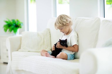 Çocuk yavru kediyle oynuyor. Siyah kediciği tutan çocuk. Küçük çocuk evdeki güneşli oturma odasında beyaz koltukta oturan sevimli evcil hayvana sarılıyor. Çocuklar evcil hayvanlarla oynar. Çocuklar ve evcil hayvanlar.