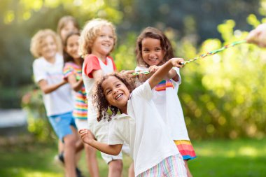 Çocuklar güneşli parkta römorkör oynuyorlar. Yazın açık hava eğlencesi. Bir grup melez çocuk spor gününde ip çekiyor. Küçük kız ve oğlan için sağlıklı bir açık hava oyunu.