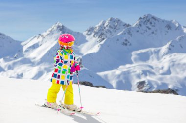 Çocuk dağlarda kayak yapıyor. Güvenlik kaskı, gözlüğü ve direkleri olan aktif bir çocuk. Küçük çocuklar için kayak yarışı. Aile için kış sporu. Alp okulunda çocuklar kayak dersi alıyor. Küçük kayakçı karda yarışıyor