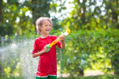 Çocuklar su püskürtme hortumuyla oynarlar. Çocuklar için yaz bahçesi eğlencesi. Sıcak ve güneşli bir günde su sıçrayan bir çocuk. Çocuk bahçedeki bitkileri suluyor. Oyuncak su tabancası.