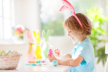 Çocuklar paskalya yumurtalarını boyuyor. Tavşan kulaklı çocuklar Paskalya avı için renkli yumurta boyuyorlar. Bahar tatili kutlaması için çiçek, sepet ve tavşanlı ev dekorasyonu. Küçük çocuk evi dekore ediyor..