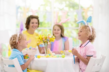 Anne ve çocuklar Paskalya yumurtalarını boyar. Anne, tavşan kulaklı küçük kız ve oğlan ölüyor ve beyaz güneşli odada Paskalya yumurtası avı için resim yapıyorlar. Aile kutlaması ve bahar tatili için ev dekorasyonu..