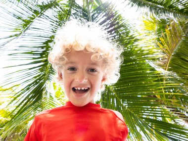 Kumsal ve yaz eğlencesi. Palmiye ağacının altındaki kıvırcık sarışın çocuk. Çocuklar deniz kıyısında oynuyorlar. Egzotik adada aile tatili. Güneşli bir günde tropikal okyanus tatil köyünde oynayan bir çocuk.. 