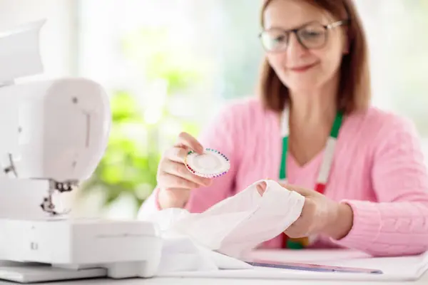stock image Woman sewing at white desk. Fashion design workshop. Creative hobby and crafts. Focus on hands. Tailor or seamstress working place with overlock machine. Female making dress. Sew equipment and tools