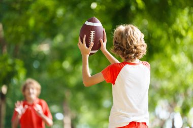 Amerikan futbolu oynayan bir aile. Çocuklar güneşli yaz parkında rugby oynuyorlar. Baba ve çocuklar top oynar, koşar ve güler. Sağlıklı açık hava sporları aktivitesi.