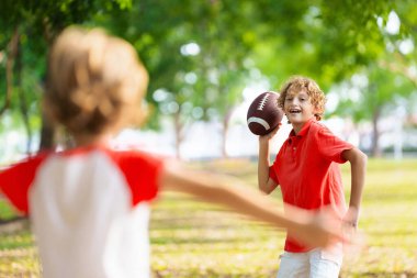 Amerikan futbolu oynayan bir aile. Çocuklar güneşli yaz parkında rugby oynuyorlar. Baba ve çocuklar top oynar, koşar ve güler. Sağlıklı açık hava sporları aktivitesi.