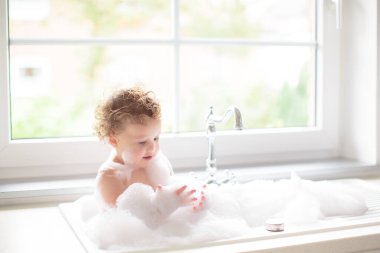 Bebek mutfak lavabosunda banyo yapıyor. Küvette sabun köpüğüyle oynayan küçük mutlu çocuk. Bir bebeği yıkamak ve yıkamak. Çocuk cilt bakımı için şampuan ve duş jeli ürünleri.