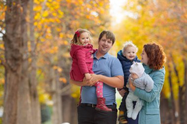 Sonbahar parkında bir aile. Ebeveynler ve çocuklar güneşli bir sonbahar gününde ormanda yürürler. Çocuklar dışarıda sarı akçaağaç yaprağıyla oynuyorlar. Küçük çocuk altın yapraklarla oynuyor. Annesi kucaklayan çocuk..
