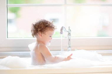 Bebek mutfak lavabosunda banyo yapıyor. Küvette sabun köpüğüyle oynayan küçük mutlu çocuk. Bir bebeği yıkamak ve yıkamak. Çocuk cilt bakımı için şampuan ve duş jeli ürünleri.