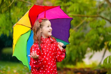 Sonbahar parkında yağmurda oynayan bir çocuk. Şemsiyeli ve çizmeli çocuk sağanak yağmurda dışarıda oynuyor. Sonbahar duşu altında kırmızı ceketli küçük kız. Çocuklar yağmurlu havada eğlenir. Çocuklar fırtınada oynuyor.