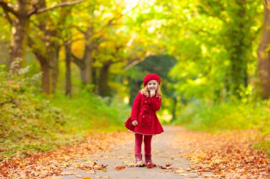Kırmızı bere ve trençkot sıcak güneşli sonbahar gününde güzel sonbahar parkı oynayan Mutlu küçük kız. Çocuklar altın akçaağaç yaprakları ile oynamak.
