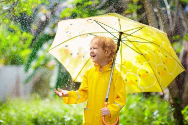 Güneşli sonbahar gününde yağmurda oynayan bir çocuk. Çocuk ağır duş altında ve sarı ördek şemsiyesiyle. Ördek su geçirmez ayakkabılı küçük çocuk. Lastik çizmeler. Yağmurlu havaya göre açık hava aktivitesi