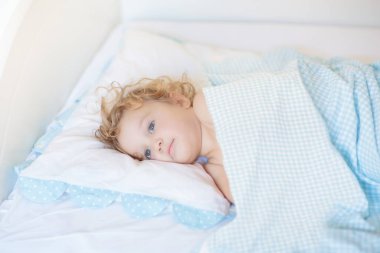 Beyaz güneşli yatak odasında sevimli bir bebek. Yeni doğmuş çocuk yatakta dinleniyor. Küçük çocuklar için çocuk odası. Çocuklar için tekstil ve yatak. Aile sabahı evde. Oyuncak ayıyla karnını doyururken doğan yeni çocuk..