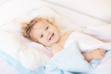 Beyaz güneşli yatak odasında sevimli bir bebek. Yeni doğmuş çocuk yatakta dinleniyor. Küçük çocuklar için çocuk odası. Çocuklar için tekstil ve yatak. Aile sabahı evde. Oyuncak ayıyla karnını doyururken doğan yeni çocuk..