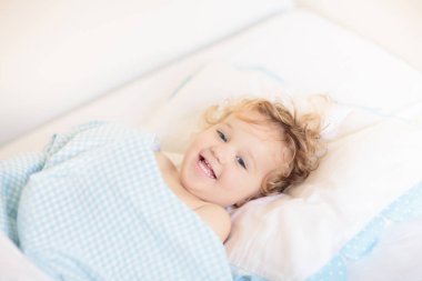 Beyaz güneşli yatak odasında sevimli bir bebek. Yeni doğmuş çocuk yatakta dinleniyor. Küçük çocuklar için çocuk odası. Çocuklar için tekstil ve yatak. Aile sabahı evde. Oyuncak ayıyla karnını doyururken doğan yeni çocuk..