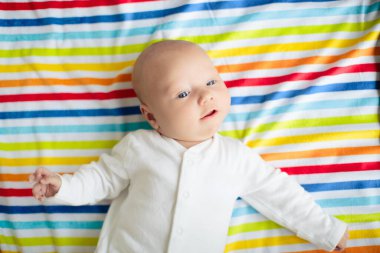 Beyaz güneşli yatak odasında, gökkuşağı battaniyesinde şirin bir bebek. Yeni doğmuş çocuk yatakta dinleniyor. Küçük çocuklar için çocuk odası. Çocuklar için tekstil ve yatak. Aile sabahı evde. Küçük çocuk için paspas oyna.