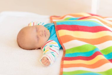 Beyaz güneşli yatak odasında sevimli bir bebek. Yeni doğmuş çocuk yatakta dinleniyor. Küçük çocuklar için çocuk odası. Çocuklar için tekstil ve yatak. Aile sabahı evde. Gökkuşağı battaniyeli yeni doğmuş çocuk.