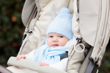 Bebek arabasındaki bebek. Kışın bebek arabasında bir çocuk. Soğuk mevsimde bebekle aktif yürüyüşler için sıcak kıyafetler. Örgü şapkalı ve ceketli şirin çocuk sandalyede oturuyor. Bebek arabasında bebek.