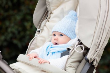 Bebek arabasındaki bebek. Kışın bebek arabasında bir çocuk. Soğuk mevsimde bebekle aktif yürüyüşler için sıcak kıyafetler. Örgü şapkalı ve ceketli şirin çocuk sandalyede oturuyor. Bebek arabasında bebek.