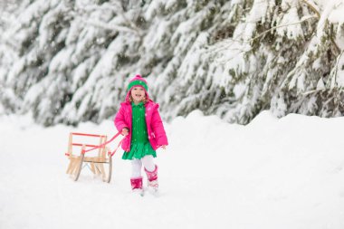 Çocuklar kışın karla oynuyorlar. Renkli ceketli küçük bir kız ve Noel 'de kış parkında kızağa binen örgü şapkalı bir kız. Çocuklar karlı ormanda kızak kullanırdı. Çocuklar için Sledge biniciliği.